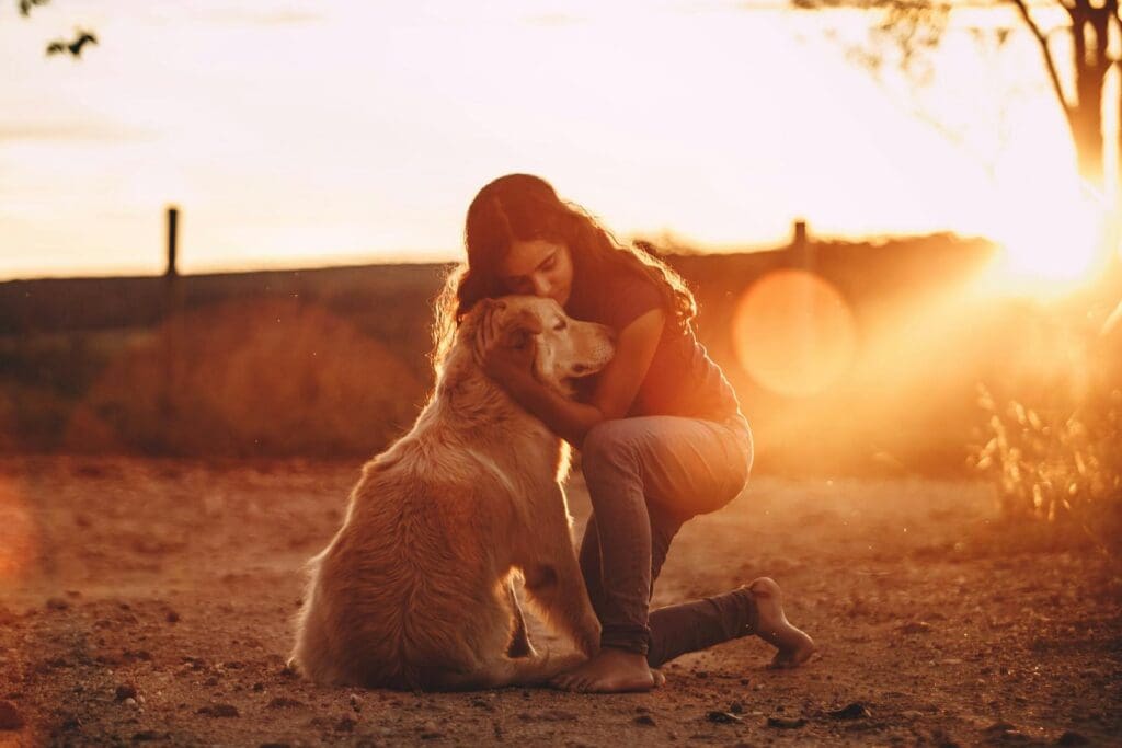 Full body of young long haired female in casual wear cuddling Golden Retriever dog while standing on knee barefoot in open air at dusk