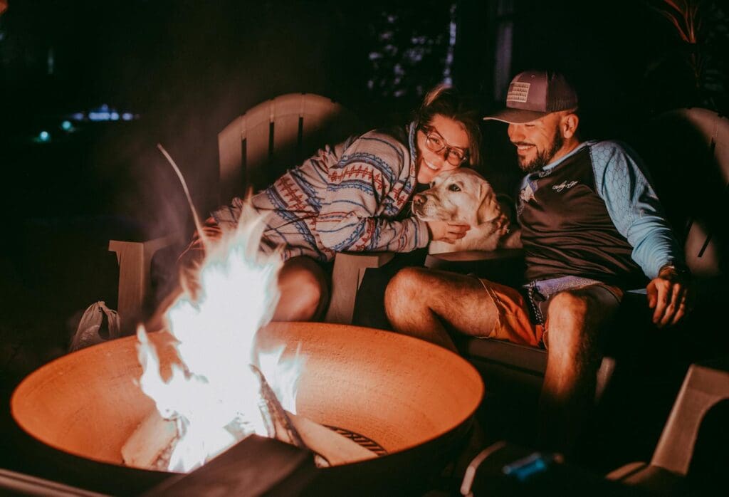 Happy young couple with Golden Retriever dog smiling and cuddling while resting near bonfire during camping in nature at night