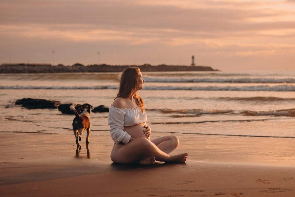 a pregnant woman sitting on a beach next to a dog
