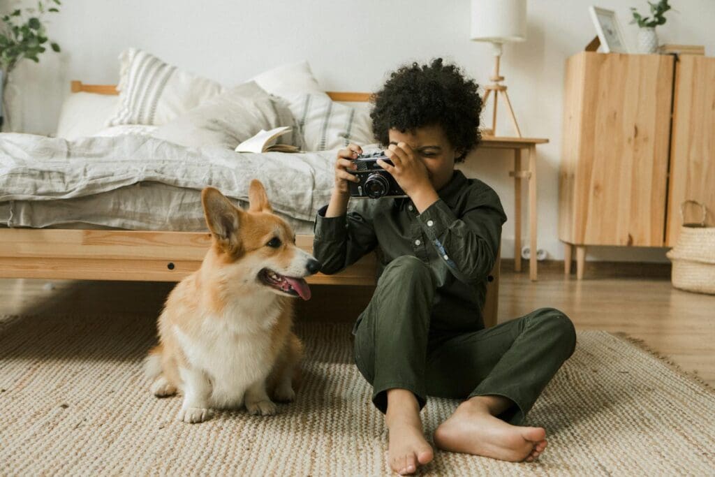 Child with camera and corgi in a cozy bedroom setting. Warm, relaxed atmosphere.