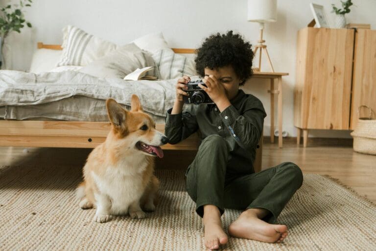 Child with camera and corgi in a cozy bedroom setting. Warm, relaxed atmosphere.