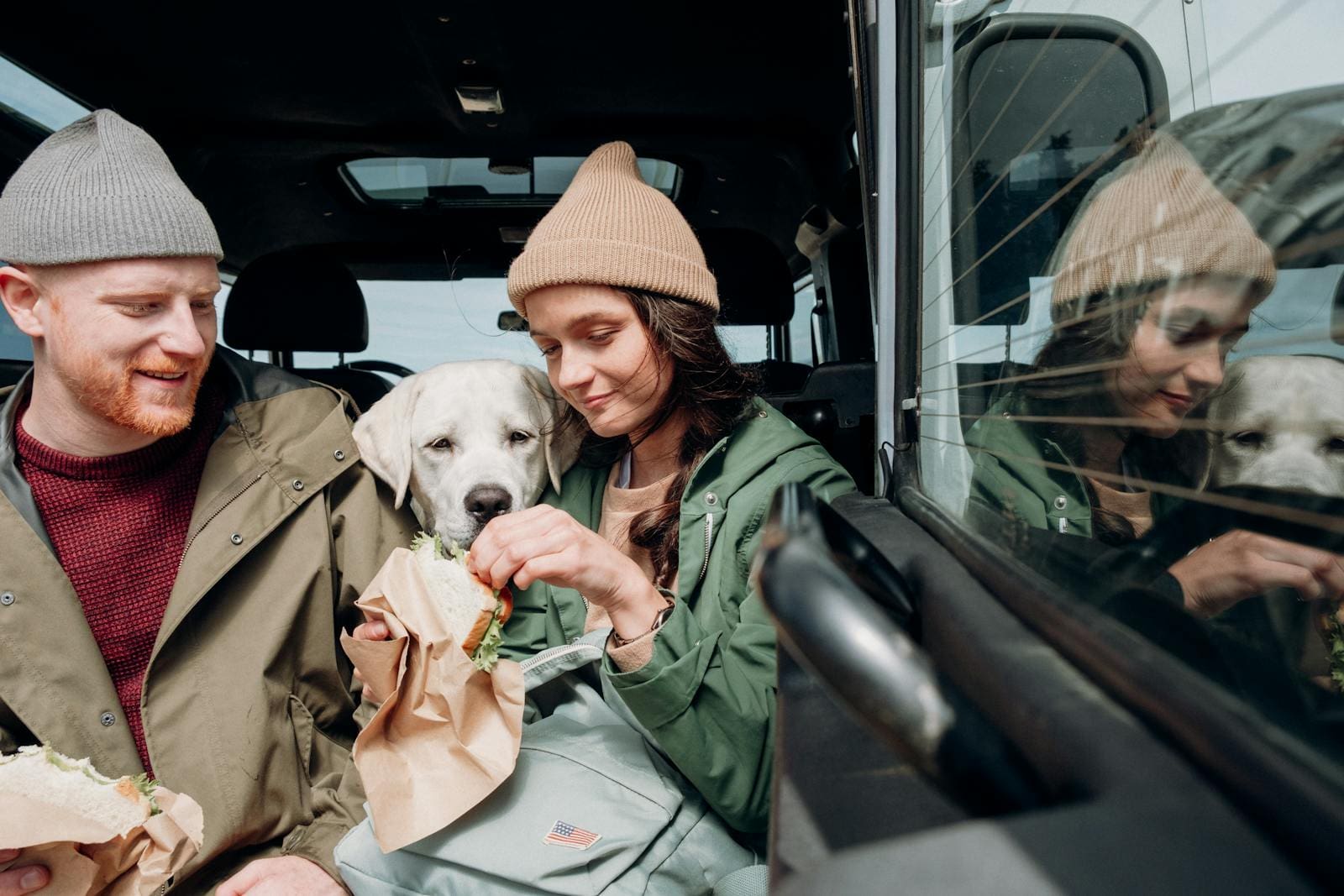 A couple with their Labrador retriever enjoying a meal inside a car on a chilly day.