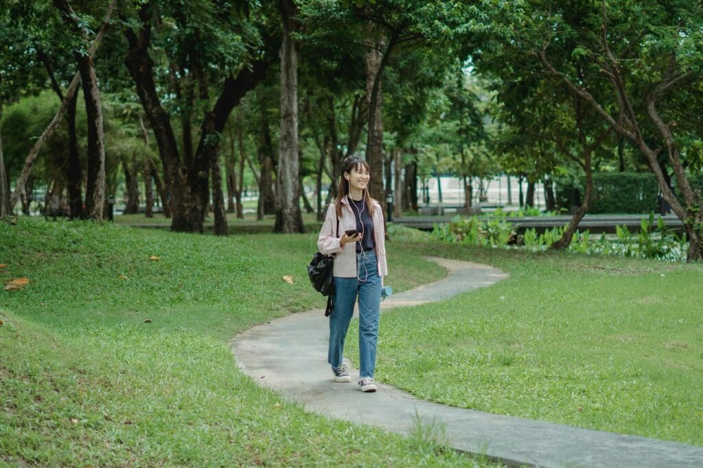 Full body of positive young Asian female in casual outfit listening to music via earphones and smartphone while walking on pathway in park