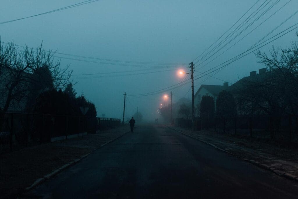 A lone person walks on a foggy street illuminated by streetlights, creating a moody atmosphere.