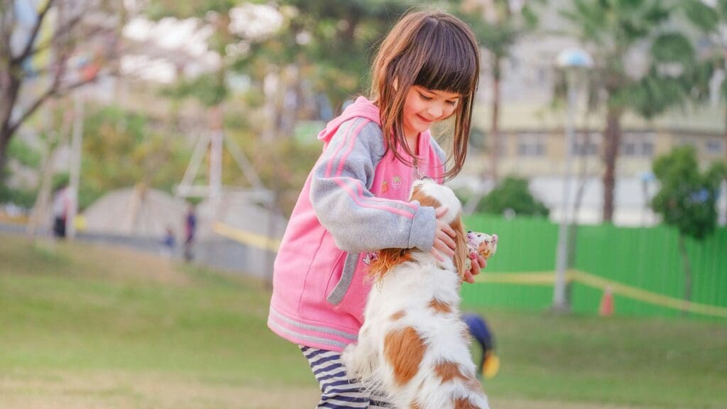 A young girl enjoys playful moments with her pet Cavalier King Charles Spaniel in a sunny park.