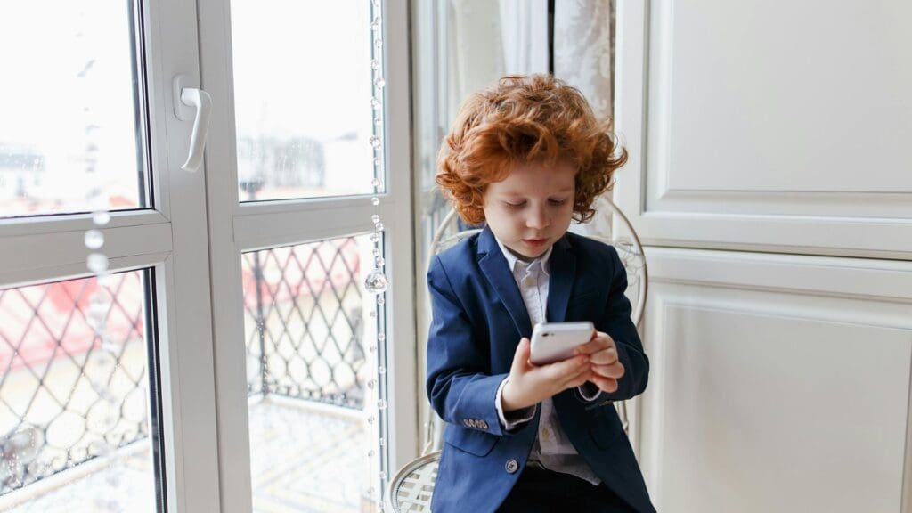 Red-haired boy wearing a suit using a smartphone indoors, seated near a window.