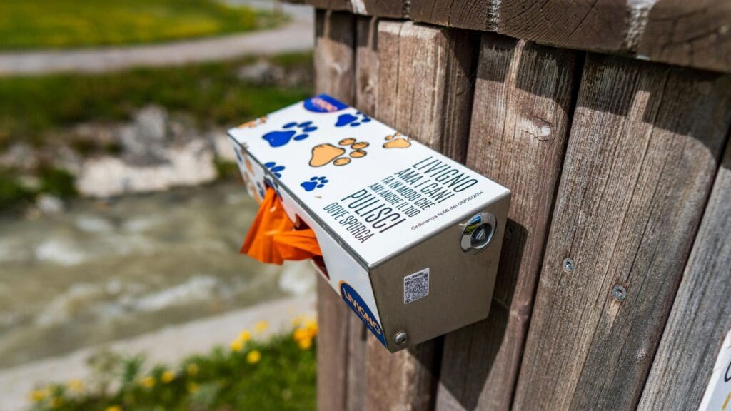 Dog waste bag dispenser mounted on wooden fence in scenic Livigno, Italy.